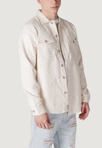 Giacca camicia beige chiaro con un motivo testurizzato, due tasche sul petto, chiusura con bottoni e colletto classico; abbinata a jeans chiari e strappati.