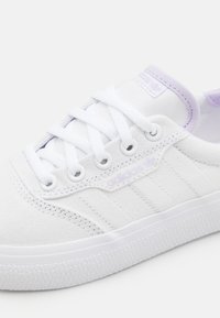 Vita canvas sneakers med en strukturerad vit sula, med lila accenter, runda snören och en logotypetikett på sidan.