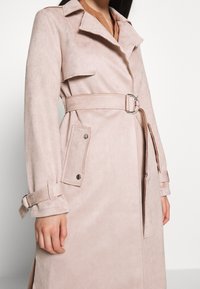 Žena v béžovém semišovém trenčku s páskem, klopovými kapsami a nastavovacími manžetami na jednobarevném bílém pozadí.