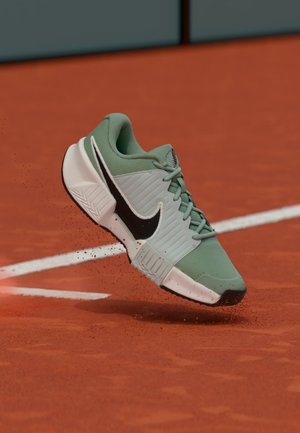 Baskets Nike vertes et blanches soulevant de la terre sur un court de tennis en terre battue rouge près des lignes blanches de délimitation.