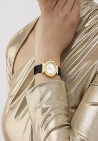 Gold-tone watch with a round face and white dial, black silicone strap, featuring stainless steel accents and a minimalist design.
