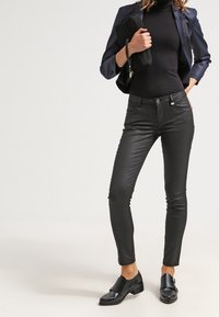 Svarta fuskläder skinny-byxor i kombination med en åtsittande svart polotröja och en marinblå blazer, hålls med en svart handväska. Svarta skor fulländar looken.