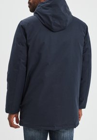 Giacca a felpa blu navy con vestibilità rilassata, texture liscia, cuciture visibili e design con schiena più lunga, indossata sopra pantaloni in denim.