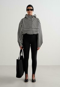 Persoon in zwarte legging en een zwart-wit houndstooth hoodie, met een zwarte hoofddoek, een zonnebril, zwarte ballerina's, en houdt een zwarte tote bag vast.