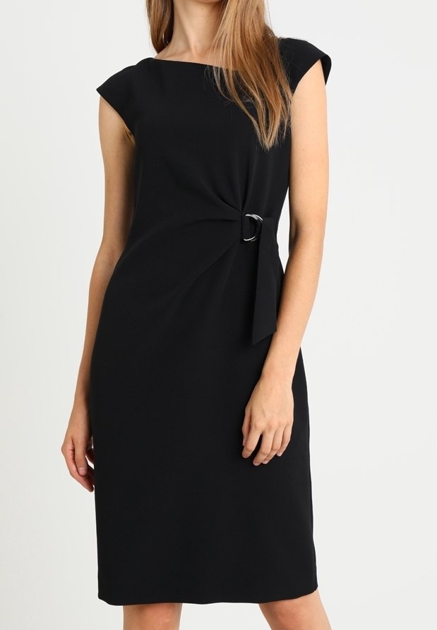 Femme portant une robe noire sans manches longueur genoux avec un décolleté bateau et une boucle métallique décorative sur le côté gauche.