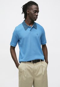 Μπλε πόλο πουκάμισο με σκούρο γιακά, υφασμάτινη υφή, κοντά μανίκια και κεντημένο λογότυπο στο στήθος, συνδυασμένο με μπεζ παντελόνια.