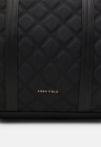 Černá prošívaná taška přes rameno s kontrastními hladkými černými popruhy. Obsahuje zlaté logo "ANNA FIELD" umístěné u dolní části.