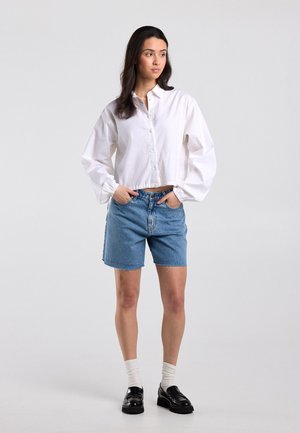 Jonge vrouw staand met handen in de zakken, draagt een witte blouse met knopen, blauwe spijkerbroekshorts, witte sokken en zwarte loafers tegen een effen grijze achtergrond.