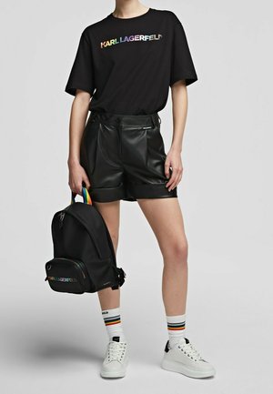 Personne portant un t-shirt noir avec un texte arc-en-ciel, un short en cuir noir, des baskets blanches, des chaussettes rayées, et tenant un sac à dos noir avec une poignée arc-en-ciel.