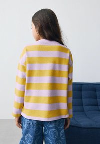 Enfant avec de longs cheveux bruns portant un pull à rayures roses et jaunes et un jean bleu, debout à l'intérieur près d'une chaise rembourrée bleue.