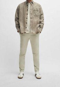 Hnědá texturovaná košile s límcem přes bílý crew neck tričko, v kombinaci s světlými béžovými kalhotami a bílými teniskami s hnědými akcenty.