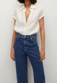 Camisa blanca de manga corta con botones, combinada con jeans de mezclilla azul de cintura alta. La camisa es suave, con puños doblados y un corte relajado.