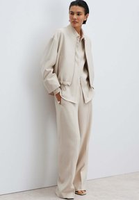 Beige cropped jacket with a drawstring hem, paired with a matching button-up shirt and wide-leg pants. Smooth fabric with a subtle sheen.