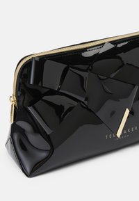 Bolso de maquillaje de charol negro con diseño de lazo geométrico, cremallera dorada y detalle de la marca en el costado. Acabado brillante, forma compacta.