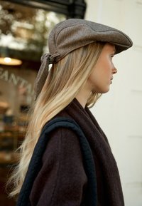 Cappello a spina di pesce marrone con un dettaglio legato sul retro, indossato da una persona con lunghi capelli lisci, abbinato a un indumento esterno scuro e strutturato.