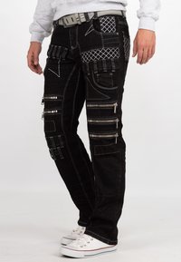 Jeans neri stile cargo con cuciture bianche, molteplici tasche con zip e dettagli testurizzati. Abbinati a sneakers bianche e una cintura.