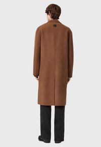 Smeđi kaput od vune s ravnim krojem, dugim rukavima i suptilnim šavom na leđima. Ima kvadratnu etiketu na kragni, nosi se preko tamnih hlača.