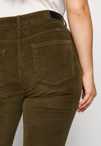 Olivenfarvede fløjlbukser med en tekstureret overflade, der viser to baglommer og et taljebånd med en sort label.