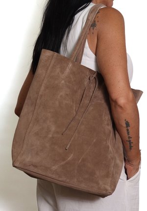 Femme vêtue d'une tenue blanche portant un grand sac cabas en suède marron sur l'épaule, montrant des tatouages sur le bras et de longs cheveux noirs.