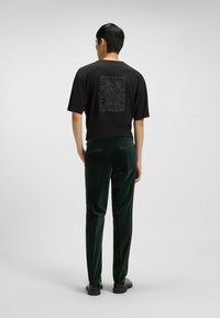 Schwarzes kurzärmliges T-Shirt mit einem strukturierten Grafikpaneel auf der Rückseite. Kombiniert mit dunkelgrünen Samenhosen. Schwarze Schuhe vervollständigen den Look.