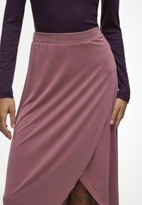 Jupe longue rose enroulée fabriquée en tissu doux avec une texture lisse. Présente une fente sur le côté et une taille élastique. Associée à un haut violet foncé.