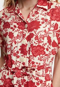 Mujer vestida con un vestido camiseros color crema con patrones florales rojos, con frutos abotonados, cuello, mangas cortas y un cinturón de lazo en la cintura.