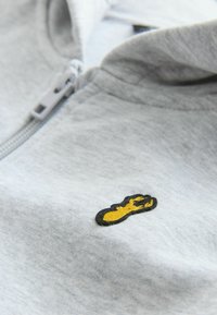 Gaiši pelēks zip-up kapuci ar dzeltenu un melnu izšūtu naglu logotipu uz krūtīm, izgatavots no mīksta, teksturēta auduma.