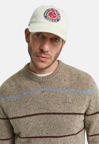 Beige Kappe mit einem roten und blauen Logo-Patch, kombiniert mit einem braunen Pullover, der blaue und burgunderrote Streifen sowie ein strukturiertes Strickmuster aufweist.