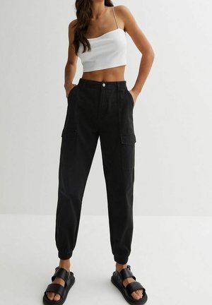 Femme portant un débardeur court blanc, un pantalon cargo noir taille haute avec des poches, et des sandales noires à bouts ouverts, debout les mains dans les poches.