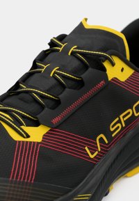 Melnas un dzeltenas sporta kurpes ar sarkanajiem šuvumiem, dzelteniem akcentu saites un "LA SPORT" zīmolu sānos.