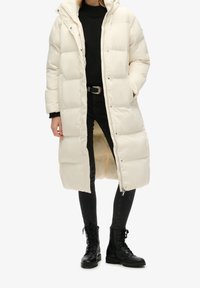 Lång, vadderad kappa i off-white med strukturerad yta, utrustad med huva, tryckknappar och sidofickor, tillsammans med svarta skinny jeans och stövlar.