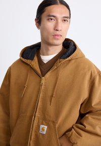 Jeune personne avec de longs cheveux sombres attachés, portant un sweat à capuche zippé marron Carhartt sur des couches de vêtements marron et blanc.