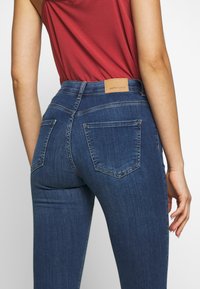 Jeans in denim blu scuro con vita alta, caratterizzati da un'etichetta in pelle marrone sul retro e due tasche posteriori con dettagli di cucitura visibili.