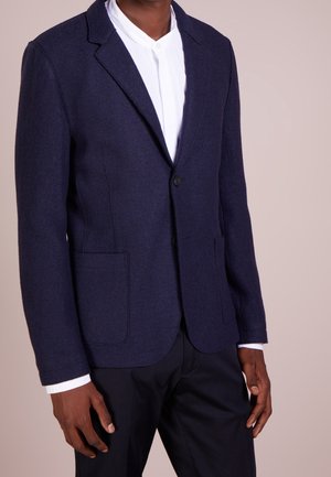 Man draagt een op maat gemaakte marineblauwe blazer over een wit overhemd en donkere broek, staand tegen een effen beige achtergrond.