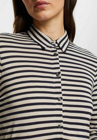 Camicia a righe con strisce orizzontali blu navy e crema, colletto e sei bottoni centrali. Realizzata in un tessuto morbido con una superficie liscia.