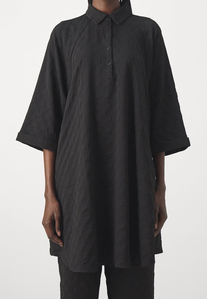 Personne portant une tunique noire texturée ample avec col et trois boutons, avec de larges manches trois-quarts et un pantalon assorti.