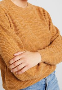 Person klädd i en texturerad senapsgul stickad tröja med långa ärmar, med armarna i kors, och ljusblå denimjeans synliga.