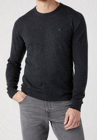 Homme portant un pull long à manches grises foncées avec des poignets et un ourlet côtelés, associé à un jean gris clair, une main dans la poche.
