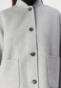 Grå feltjakke med ståkrave, der har fire store knapper og to frontlommer. Glat tekstur og struktureret design.