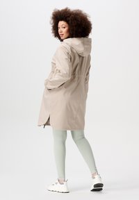 Beige vattentät jacka med huva, åtdragen midja och sidofickor. Bärs med ljusgröna leggings och vita sneakers. Slät textur.