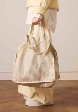 Beige textuur tote bag met twee stevige handvatten, met een gestreept patroon en zacht materiaal. Geplaatst tegen een lichte achtergrond.