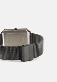 Montre en acier inoxydable avec un bracelet en maille noire mate, un boîtier rectangulaire et des détails argentés polis ; elle possède un fermoir à clapet.