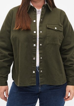 Personne portant une veste vert olive déboutonnée sur une chemise blanche à col et un jean bleu, cadrée au-dessus des épaules et à mi-cuisse.