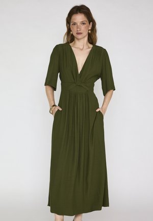 Femme portant une longue robe vert olive avec un profond décolleté en V, des manches mi-longues et des poches latérales, debout devant un fond uni.
