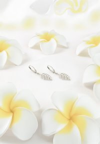 Sølv bladeformede øreringe med stribet detaljering placeret blandt hvide og gule blomsteraccenter på en blød baggrund.