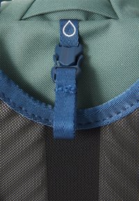Tejido verde con un acabado texturizado, que presenta una correa azul con hebilla y un logo de gota blanca encima. Se pueden ver las costuras acentuadas.