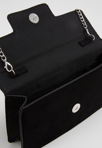 Bolso rectangular negro con tapa abierta, cierre de botón de presión plateado, bolsillo interior con cremallera y correa de cadena plateada con cierre tipo langosta.