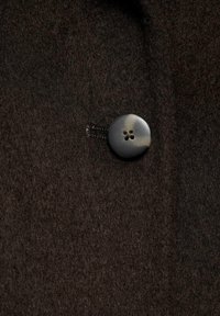 Gros plan sur un manteau en laine marron foncé avec un unique bouton noir rond à quatre trous fermé à travers une boutonnière cousue.