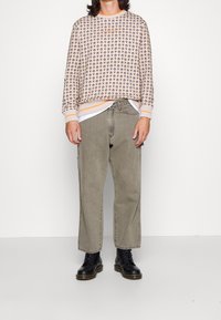 Maglione beige con motivi marroni scuri ripetuti, dettagli arancioni nel colletto e nei polsini. Jeans baggy verde oliva e stivaletti neri con lacci.