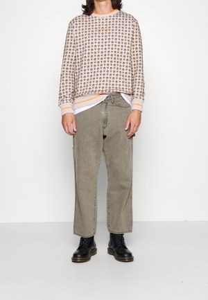 Maglione beige con motivi marroni scuri ripetuti, dettagli arancioni nel colletto e nei polsini. Jeans baggy verde oliva e stivaletti neri con lacci.